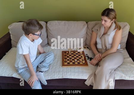 Mother and son playing chess on a beige couch. A woman and a boy sit on a beige couch in front of a green wall, concentrating on a chess game. Stock Photo