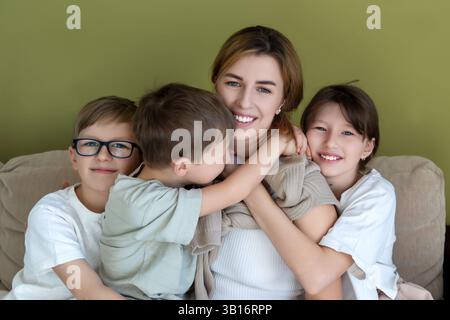 Happy mother embracing her three children at home. Smiling mother sitting on a couch, surrounded by her three children in a warm embrace. A loving and Stock Photo