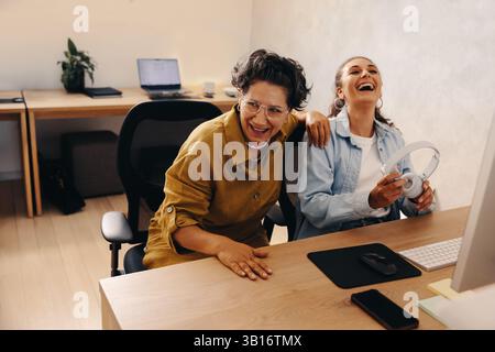 Two coworkers share a joyful moment while seated at a desk in a modern workplace. Their laughter reflects a positive work environment and a sense of c Stock Photo