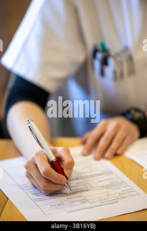Female healthcare professional focuses on writing notes at desk, surrounded by pens and medical equipment, indicating busy clinical environment Stock Photo