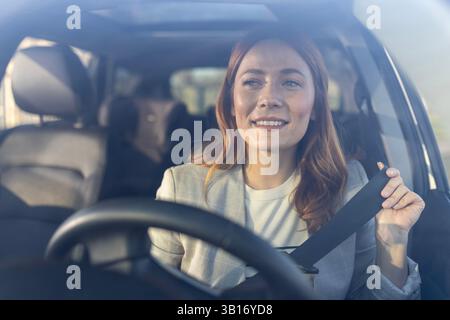 Beautiful business woman with red hair in drivers seat smiling as she puts on her safety seatbelt before driving a car Stock Photo