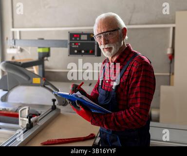 Skilled older man inspects plans while working in woodworking shop, wearing safety glasses and gloves, showing dedication to his craft. Stock Photo