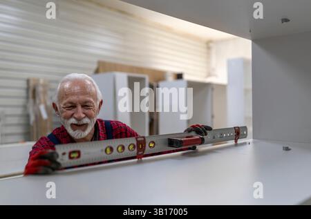 Senior man carpenter in red plaid shirt uses a level to ensure the accuracy of a shelf installation in a well-lit workshop Stock Photo