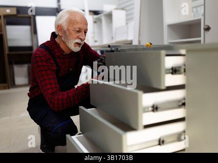 An elderly man with a beard focuses on assembling set of drawers in well-lit workshop, showcasing craftsmanship and attention to detail during the tas Stock Photo