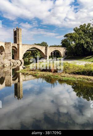 A medieval stone bridge with arches reflecting in a calm river, surrounded by lush greenery and a partly cloudy sky. Stock Photo