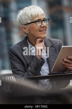 Smiling mature business woman looking away from tablet and thinking while sitting outdoors Stock Photo