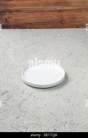 Minimalist white ceramic plate placed on a textured concrete surface with wooden backdrop. Ideal for food photography, tableware mockups, or clean mod Stock Photo