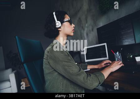 Young female coder working in a modern office environment with programming code on computers, wearing headphones, focused on programming Stock Photo