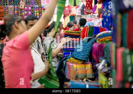 Zegyo Market Colorful Stacked Longyi Fabrics Mandalay Myanmar | MANDALAY, Myanmar — A shopper sorts through stacks of brightly patterned longyi fabric at a crowded textile stall in Zegyo Market. The zigzag acheik motifs visible on several folded pieces trace back to weaving patterns developed for the nineteenth-century Mandalay royal court. Textiles by the bolt, including silk, cotton, and synthetic cloth, remain among the principal goods sold at Zegyo, Mandalay's central market since King Mindon founded the city in the mid-1850s. Stock Photo