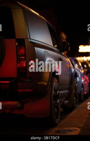 A matte gray Land Rover Defender parked on a city street at night, tail lights glowing red, with blurred streetlights in the background. Stock Photo