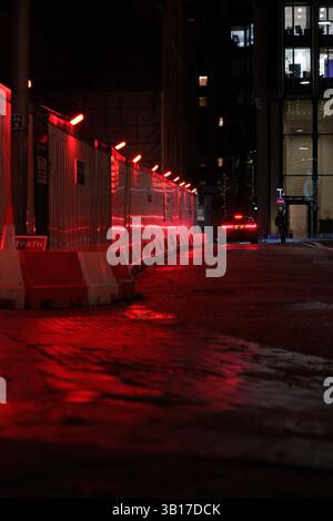 Red lights illuminate a city street at night, casting a glow on construction hoardings and a parked car. Stock Photo