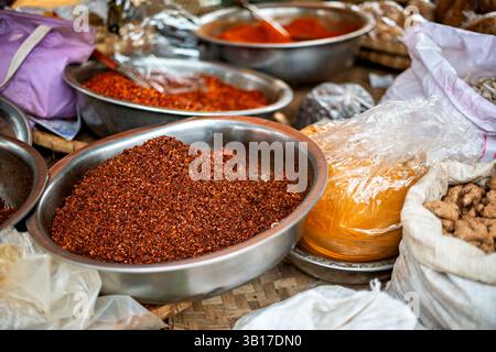 Chili Powder At Zegyo Market Mandalay Myanmar | MANDALAY, Myanmar — Bowls of ground chili powder and other spices fill a vendor's stall in the street market surrounding Zegyo Market, Mandalay's principal central bazaar since the city's founding under King Mindon in the 1850s. Sold loose by weight, chili powder is a foundation ingredient in Burmese cooking, giving curries and relishes their color and heat. Vendors here often process their own chilies, and the stalls reflect Mandalay's long history as a crossroads for spices moving between India, China, and the Irrawaddy valley. Stock Photo