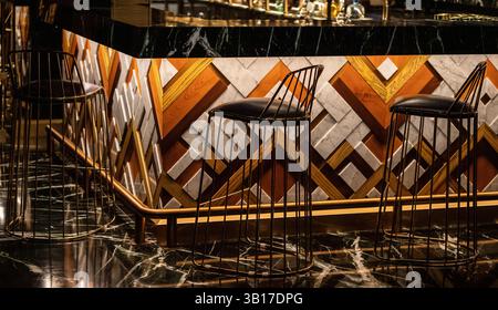 Stylish bar counter with geometric wood and marble paneling, illuminated by warm lighting, featuring modern wire stools and a dark marble floor. Stock Photo