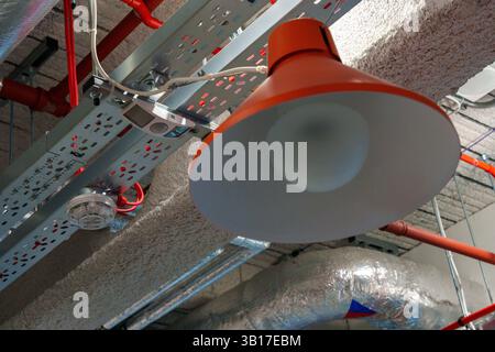 Low angle view of an orange industrial lamp, fire alarm, motion sensor, and exposed pipes and wiring in a modern building. Stock Photo