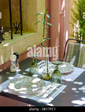 A charming al fresco dining setting bathed in dappled sunlight, featuring floral plates, green glassware, and a lucky bamboo centerpiece in Greece. Stock Photo