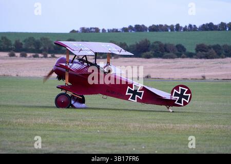 A historic Fokker Dr.I triplane, painted red with black Iron Cross markings, sits on a grassy airfield, propeller blurred at Duxford, England. Stock Photo