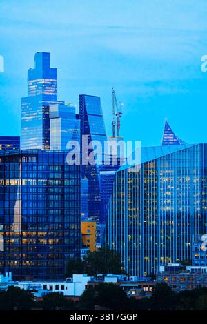 Modern skyscrapers illuminate the twilight sky in the City of London financial district, England, with a crane hinting at ongoing development. Stock Photo