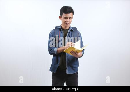 Asian man wearing dark blue denim shirt carrying backpack and open book studying for exam preparation, isolated on white background Stock Photo