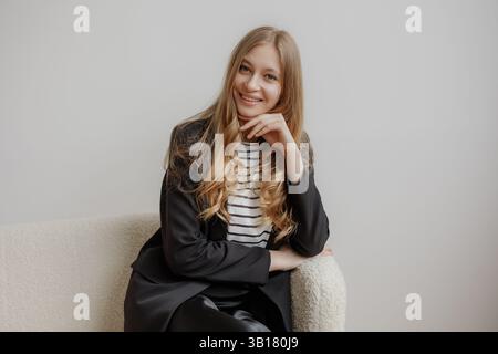 Young woman with long hair wearing a striped shirt and blazer poses confidently in a cozy indoor setting Stock Photo