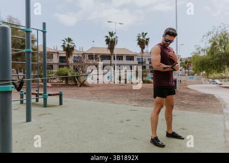 A fit man is training at an outdoor gym using a fitness band . He is wearing a red shirt and black shorts Stock Photo