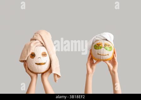 Female hands holding pumpkins with drawn faces, clay masks and towels on light background Stock Photo