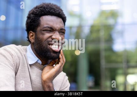 Man suffering from toothache. African American businessman sitting on a bench outside an office building and holding his hand over his gap, inflammation of the gums. Stock Photo