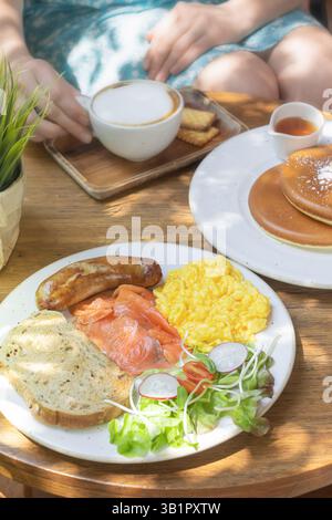 A beautifully arranged breakfast includes scrambled eggs, smoked salmon, a sausage link, salad, and artisanal bread. On the side, a stack of fluffy pa Stock Photo