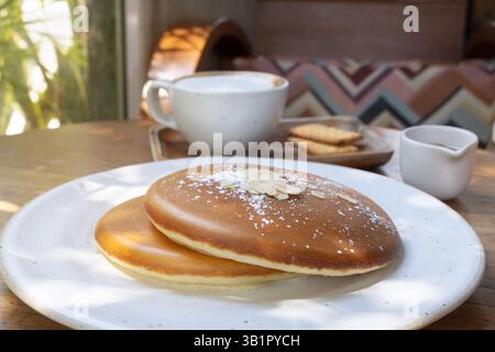 A cozy In the morning, at a sunny cafe. A stack of fluffy homemade pancakes sits on a white plate, topped with a drizzle of sweet syrup. Served alongs Stock Photo