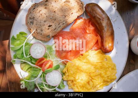 A hearty breakfast platter showcases a vibrant assortment of smoked salmon, fluffy scrambled eggs, a juicy sausage, and fresh vegetables including lea Stock Photo
