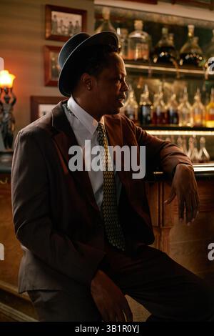 Side view of stylish African American male jazz vocalist attired in vintage hat and suit sitting at high top table near bar, illuminated by soft dim light creating intimate ambience Stock Photo