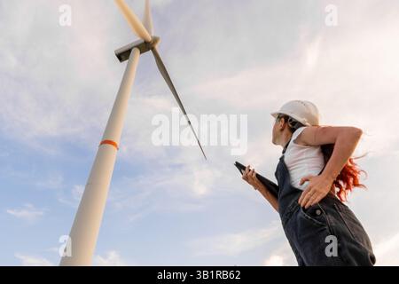 A young female engineer studies a wind turbines design, analyzing its functions and harnessing renewable energy under a vibrant, cloudfilled sky that fuels her sustainability commitment Stock Photo
