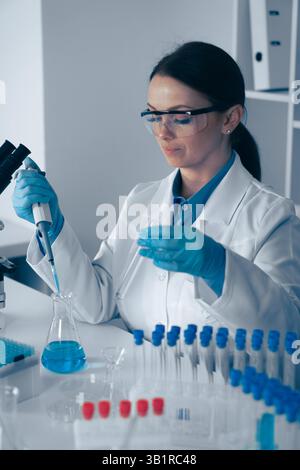 A focused female scientist in a lab coat and gloves measures a blue liquid with a pipette, surrounded by test tubes and lab equipment, conducting essential experiments for scientific advancement Stock Photo