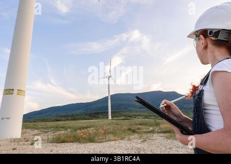 A highly skilled female engineer stands confidently alongside a large wind turbine model, demonstrating her unwavering commitment to promoting renewable energy and sustainability at a local wind farm Stock Photo
