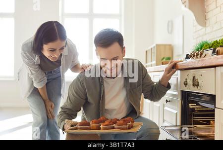 Portrait of young happy couple looking at baked muffins on baking tray standing in kitchen. Stock Photo