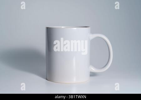 A plain white ceramic mug with handle standing empty on a smooth light gray surface, captured in soft lighting with minimal shadows. Stock Photo