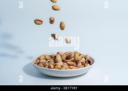 Pistachio nuts falling into a white ceramic bowl already filled with pistachios, captured against a clean light blue background. Stock Photo