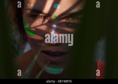Detailed close up of beautiful woman with neon creative painting make up showing her face through palm leaves. Fashion model with shadow on her face. Stock Photo
