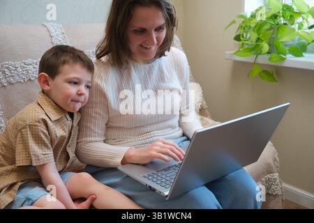 Mother and son sitting on a couch using a laptop at home. Family bonding and online learning concept. Stock Photo