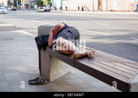 Unrecognizable barefoot homeless man sleeps on a bench on a city street, homelessness, social issues Stock Photo