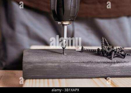 Repairman screwing with electric screwdriver black screw into a wooden plank, carpentry, DIY repair Stock Photo