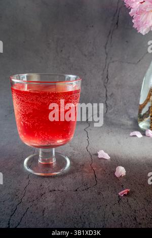 A glass of red drink with bubbles in it sits on a table next to a vase of pink flowers. grey background Stock Photo