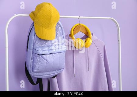 Stylish lilac hoodie with headphones, backpack and cap on rack against color background Stock Photo