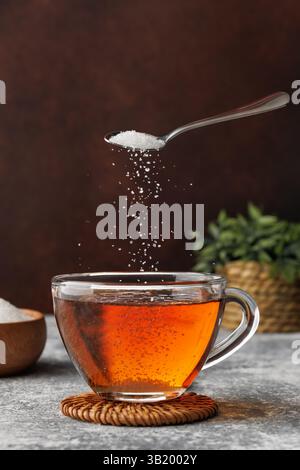 White sugar falling from teaspoon into glass cup of hot tea placed on coaster against dark brown background Stock Photo