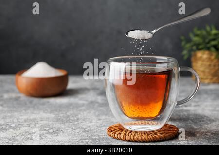 White sugar falling from teaspoon into glass cup of hot tea placed on coaster against grey background Stock Photo
