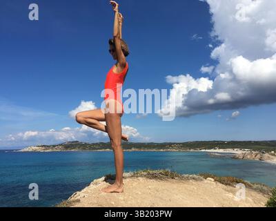 Beautiful sporty woman wearing orange swimsuit doing active pose with one leg up  on the edge of sea view point in Italy in Sardinia. Stock Photo