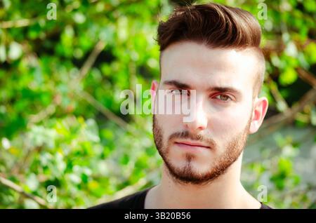 A young man stands confidently in front of lush green foliage. His well-groomed beard and trendy hairstyle add to his modern appearance as he gazes thoughtfully into the distance. Stock Photo