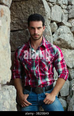A young man stands with a serious expression, dressed in a plaid shirt and jeans, leaning against a rough stone wall. The natural setting provides a rustic backdrop. Stock Photo