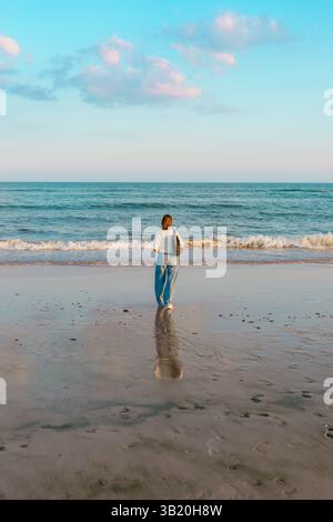 Woman facing the ocean, standing at the water's edge with her reflection visible on the wet sand. Stock Photo