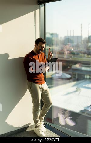 A man engages with a tablet while smiling, standing by a large window in a contemporary office. The bright sunlight highlights his thoughtful expressi Stock Photo