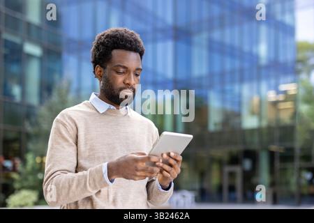 A focused African American man reads or works on his tablet outdoors, with a modern office building in the background. The natural light enhances the scene. Stock Photo